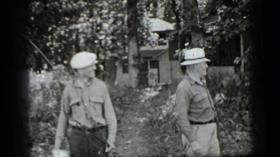 Two men stand in a shaded Clear Lake cabin area, one looking down with a fishing rod and bucket nearby.
