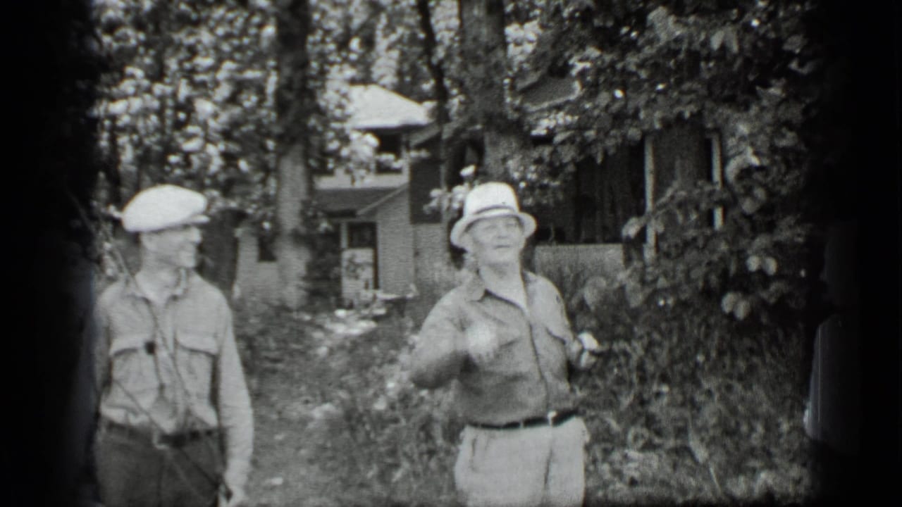 Two adults in hats stand in a leafy garden with a house behind them.