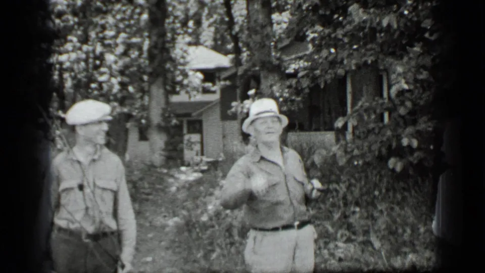 In 1939 Clear Lake home-movie footage, two adults in hats stand among trees and foliage with a house partly visible behind them.