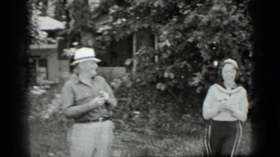 A man in a brimmed hat stands opposite a woman in a sailor-collar sweater in the Clear Lake garden setting.
