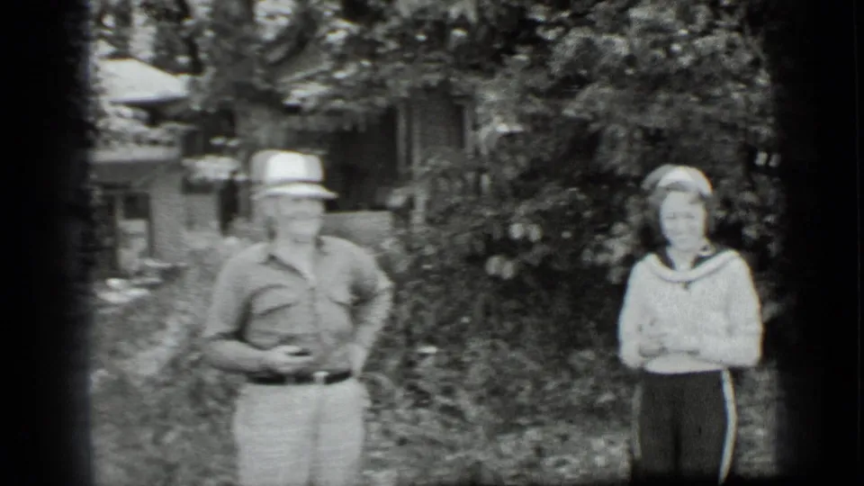 A man and woman stand in a leafy Clear Lake yard, with soft-focus 8mm texture and a house partly visible beyond the foliage.