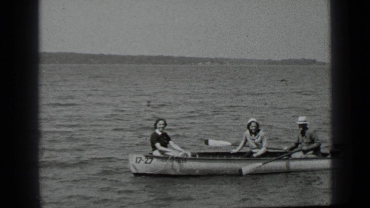 Three people sit in a small rowboat on a lake with oars visible.