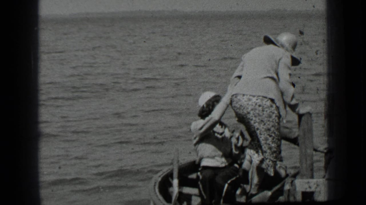 A woman in a hat helps someone near a small boat beside a dock.