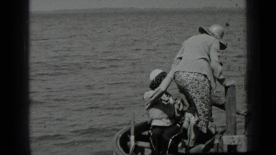 At Clear Lake in 1939, a woman in a wide-brimmed hat and patterned skirt appears to help another person into a small boat by the dock.