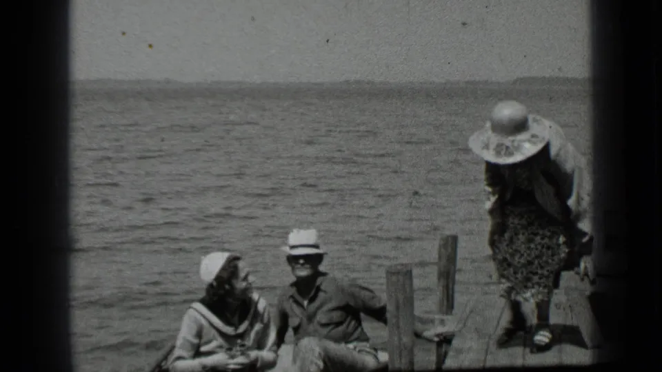 Clear Lake dock scene from 1939 with two adults seated by the water and a woman in a hat standing near the dock edge.