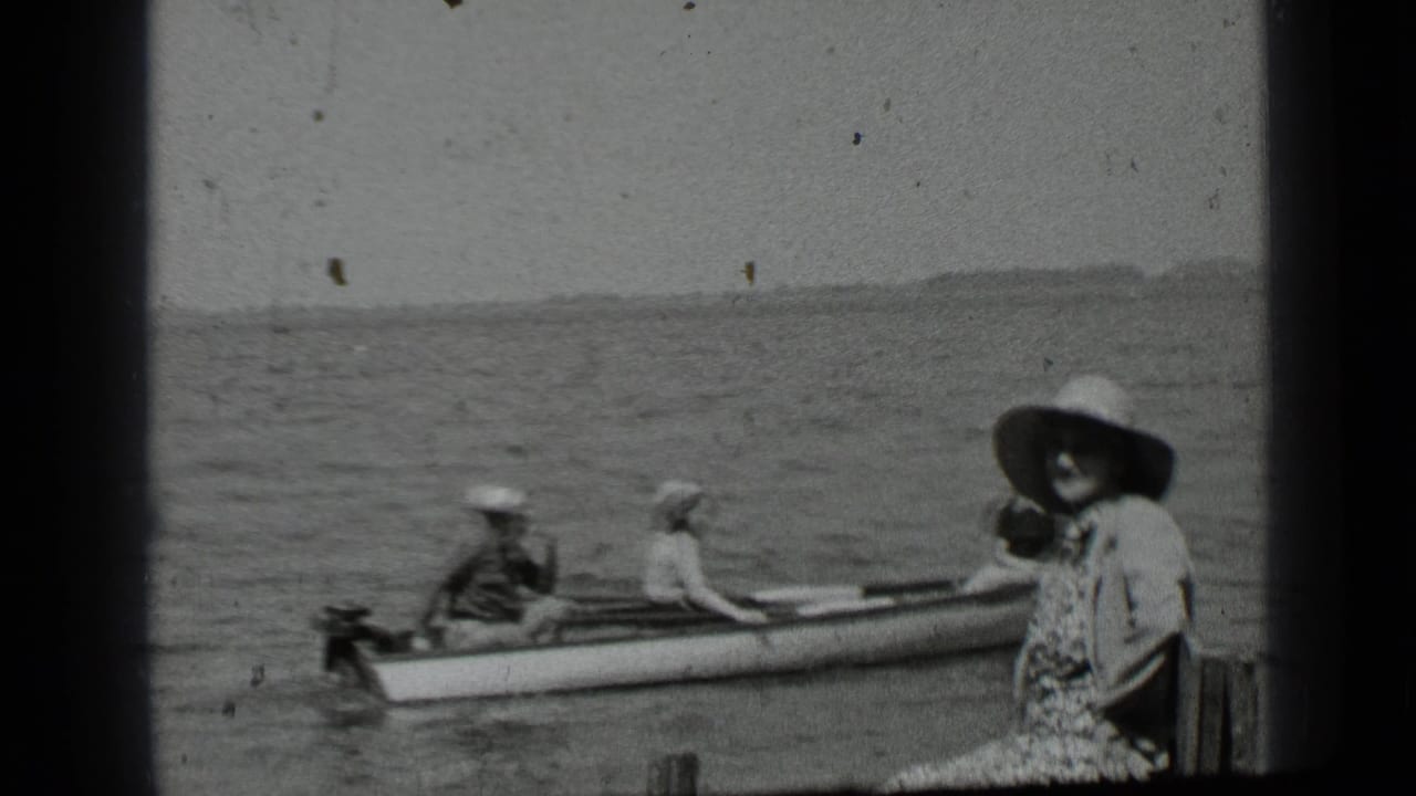 People in hats sit in a small boat beside a dock on a lake.