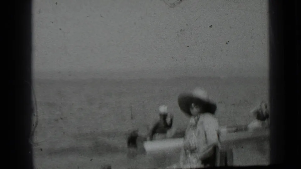 A soft 8mm frame shows a small boat on Clear Lake moving near a seated woman in a wide-brimmed hat by the water.