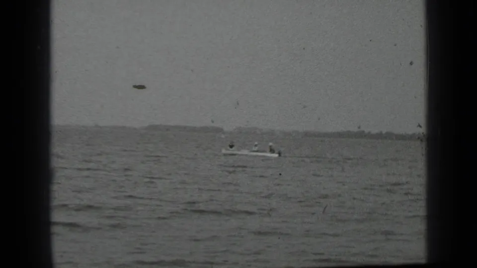 A small boat appears on Clear Lake, California, with people faintly visible aboard and a low shoreline under a gray sky.
