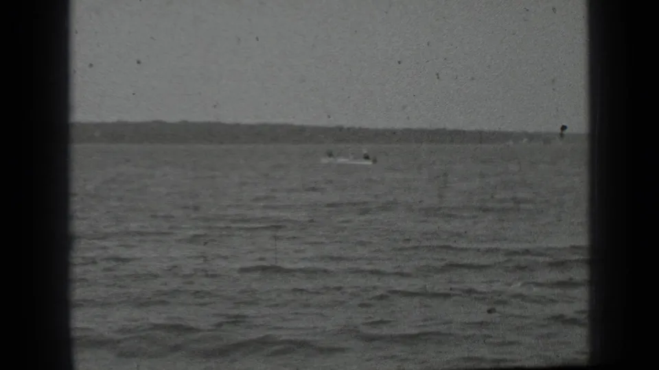 Home-movie frame of a distant small boat on Clear Lake, with choppy water filling the foreground and land along the horizon.