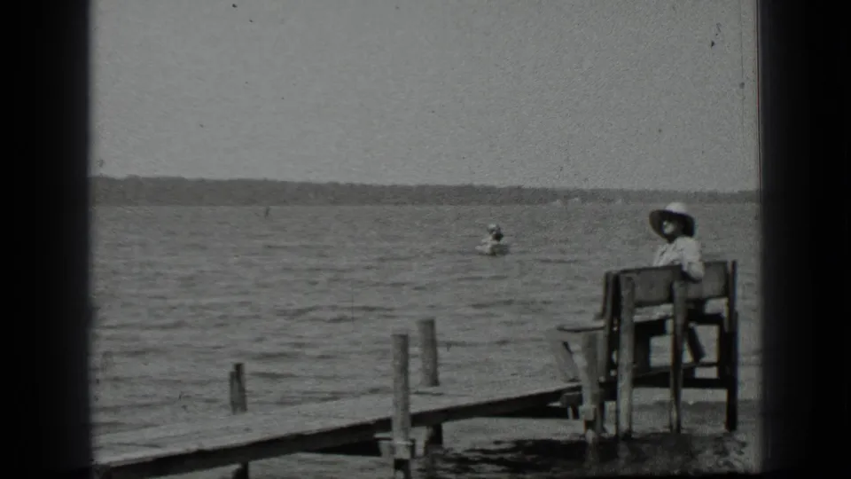 1939 Clear Lake home-movie frame showing a woman in a wide-brimmed hat seated by the dock while a canoe is visible on the water.