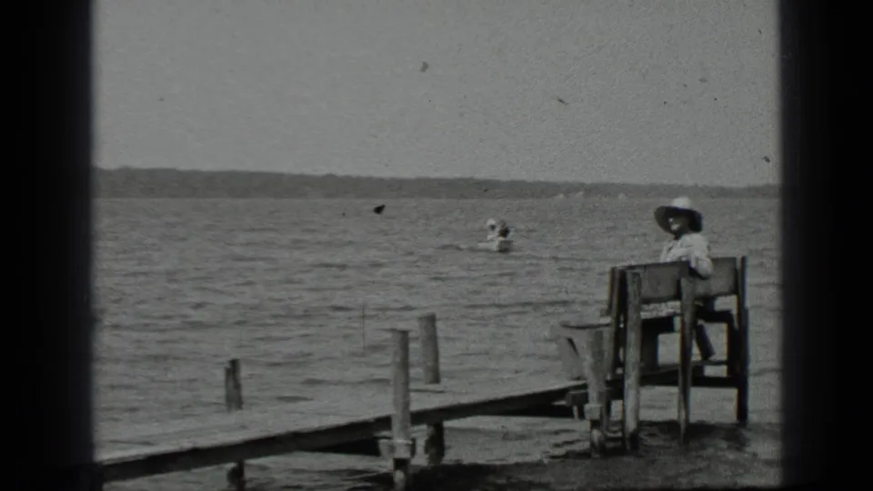 At Clear Lake in 1939, a woman sits on a wooden bench near the dock while a small canoe appears beyond her on the rippled water.