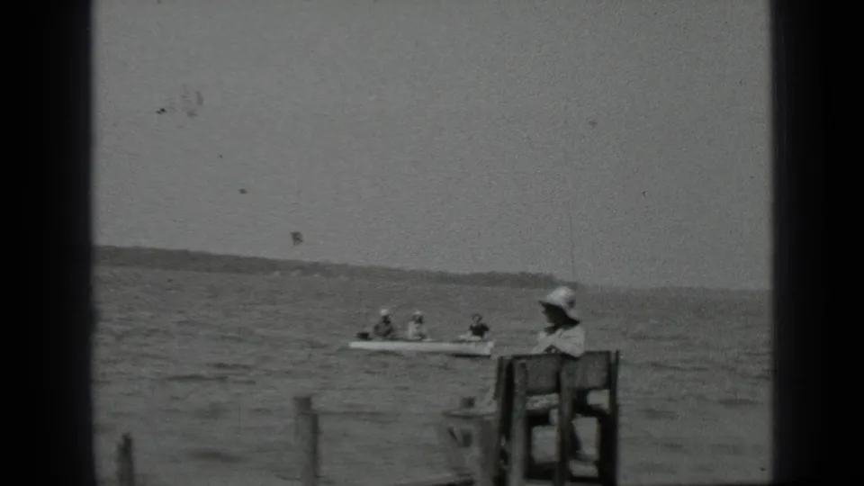 1939 Clear Lake footage shows a canoe with several people passing closer to the dock where a woman sits on a wooden bench.