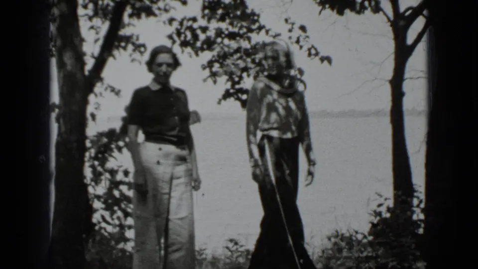 Two women stand beside trees at Clear Lake, California, one in light trousers and one in darker trousers, with water behind them.
