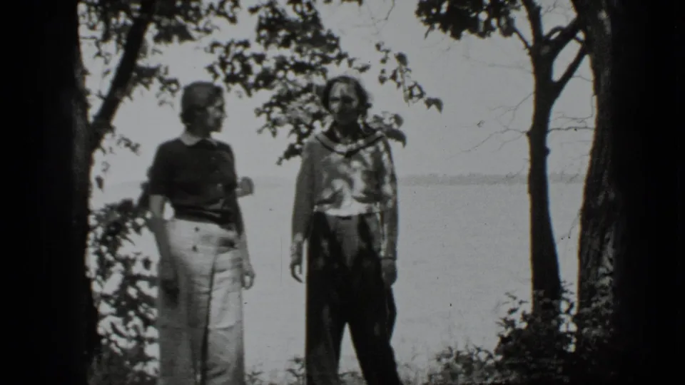 Black-and-white 8mm frame of two women posing near trees at Clear Lake with the lake visible through the background.
