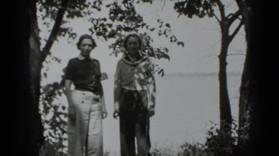 Two women face the home-movie camera at the Clear Lake shore, framed by trees and bright water in the background.