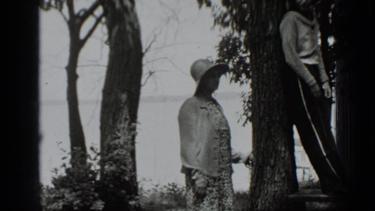 Girl in a brimmed hat stands by trees while a boy leans nearby with water in the background.