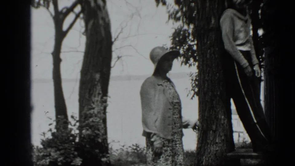 1939 Clear Lake home-movie frame showing a girl in a brimmed hat and patterned dress near trees as a boy leans beside her.