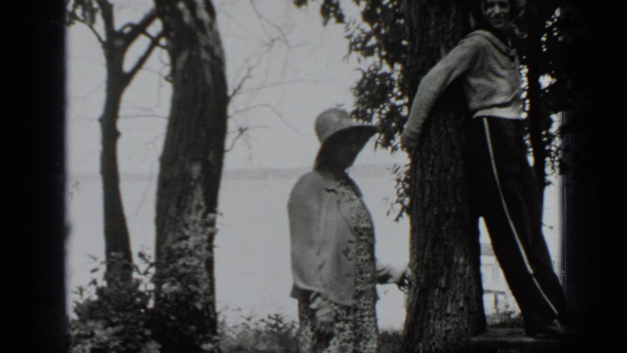 Girl in hat faces a boy leaning against a tree near shrubs and a lake.