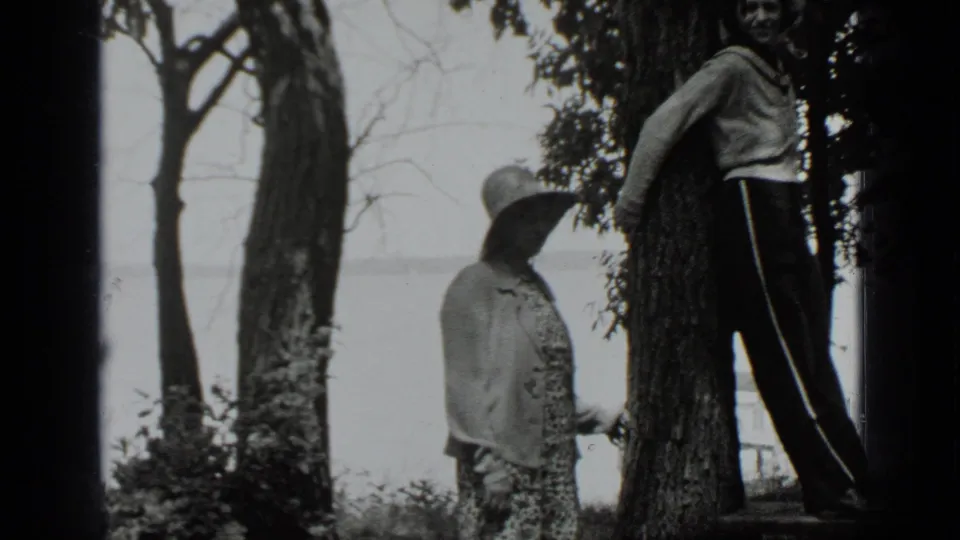 1939 Clear Lake frame with a girl in a wide-brimmed hat facing a boy leaning by a tree in a wooded lakeside setting.