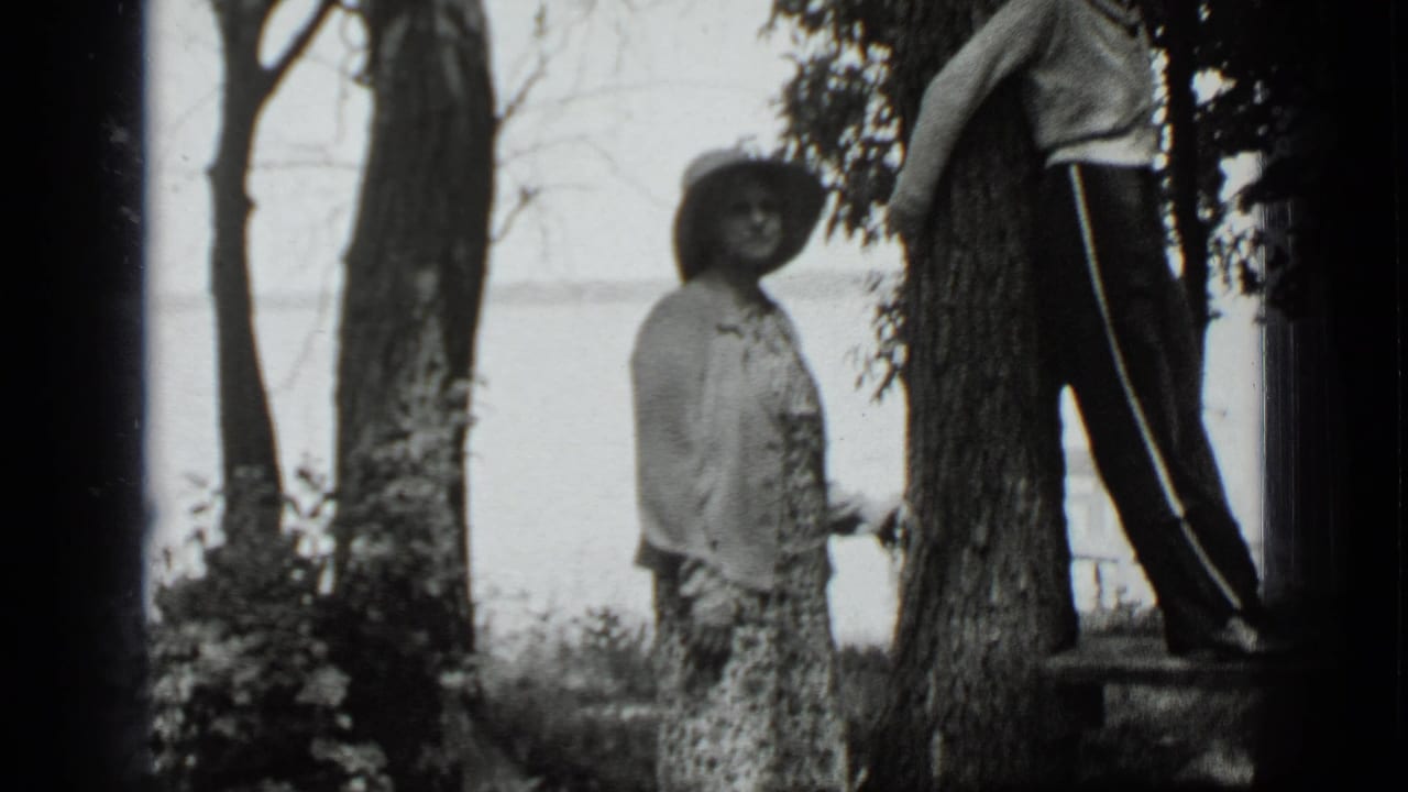 Two adult women stand among trees near a lake, one in a wide-brimmed hat and floral dress.