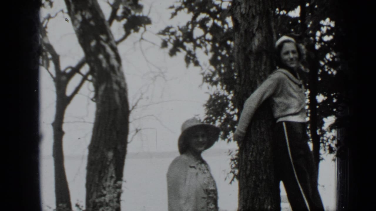 Two adult women stand by trees with lake water behind them, one partly cropped at right.