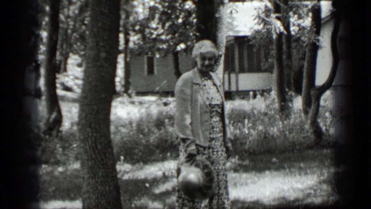 Woman standing among trees near a house, holding a hat at her side.