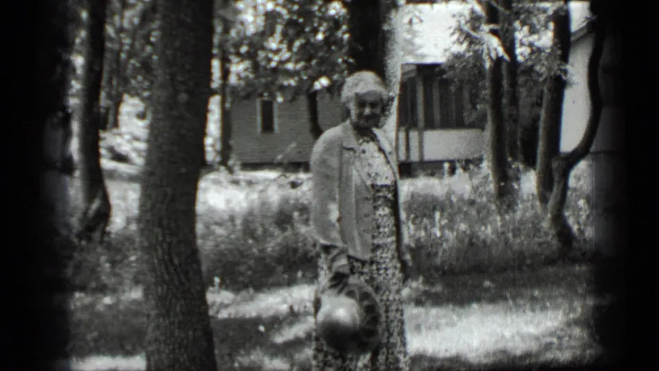 In 1939 Clear Lake home-movie footage, a woman in a patterned dress and light jacket stands in a wooded yard holding a hat.