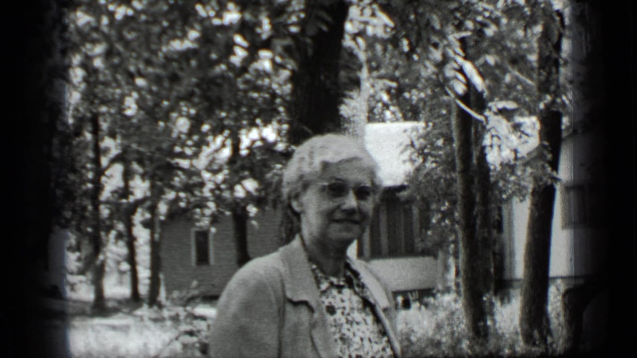 Older woman in glasses stands in a leafy yard with a house behind her.