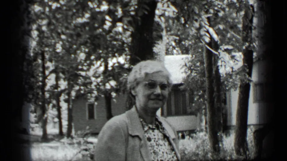 Older woman in glasses and a cardigan stands outdoors in Clear Lake, California, with trees and a modest house behind her.