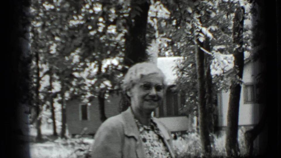 Smiling older woman in a cardigan appears in a leafy Clear Lake yard, with a house exterior partly visible behind her.
