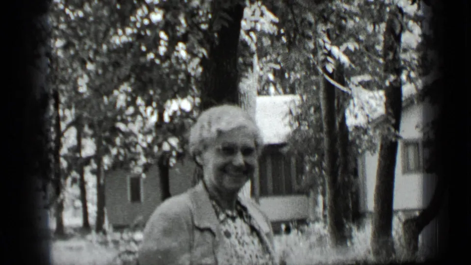 Older woman in glasses and a light cardigan smiles at the 8mm camera in a leafy Clear Lake residential yard.