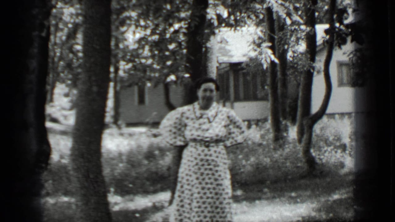 Woman in a polka-dot dress standing among trees near a house.
