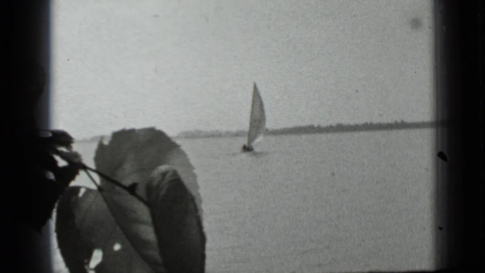 A small sailboat moves across Clear Lake in a black-and-white 8mm view partly framed by foreground leaves.