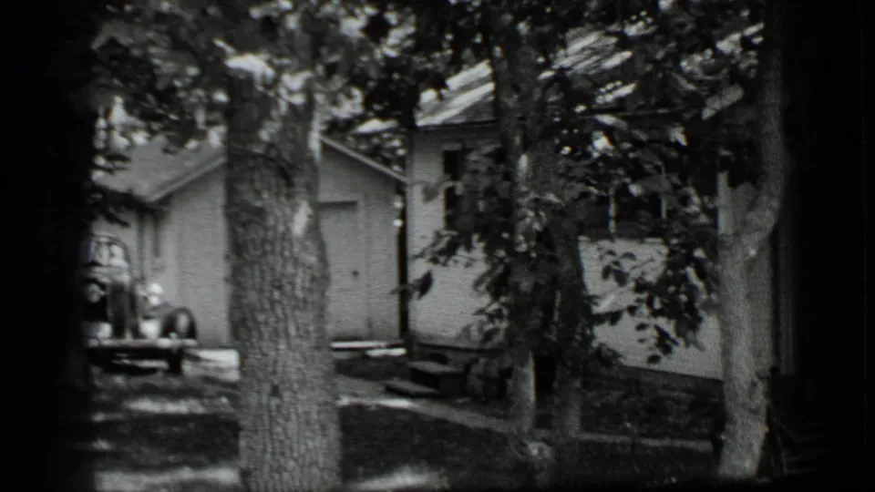 1939 Clear Lake home-movie frame showing a white house and small shed behind large trees in a shaded yard.