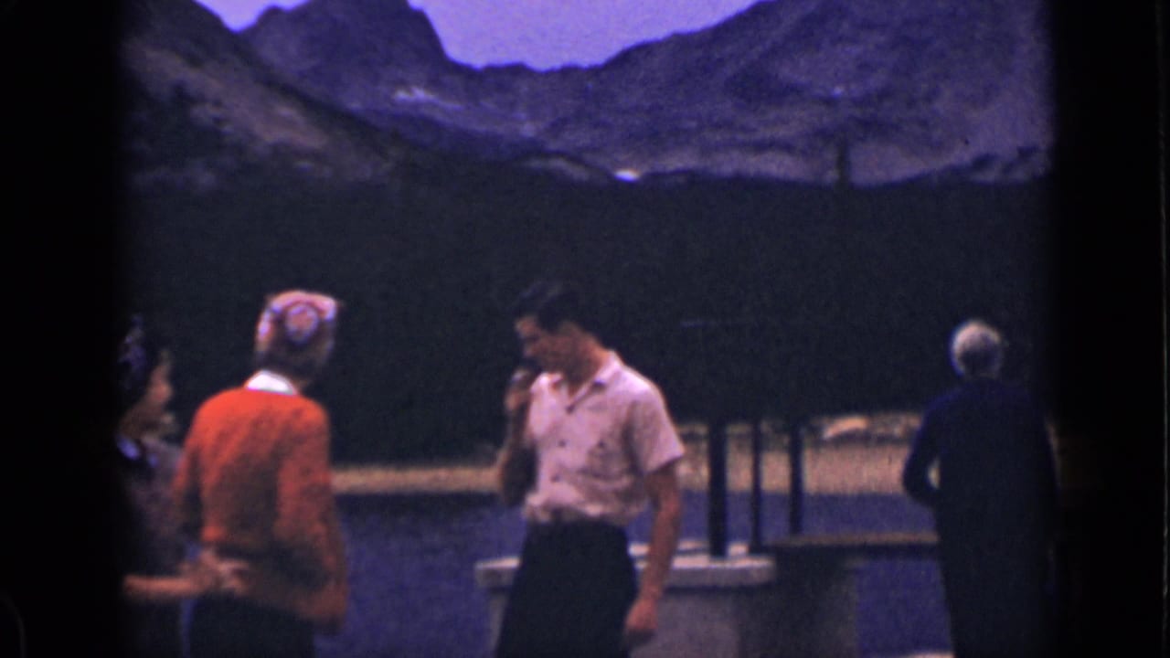 In 1939 Colorado home-movie footage, adults gather by a stone bridge with lake water, trees, and mountains behind them.