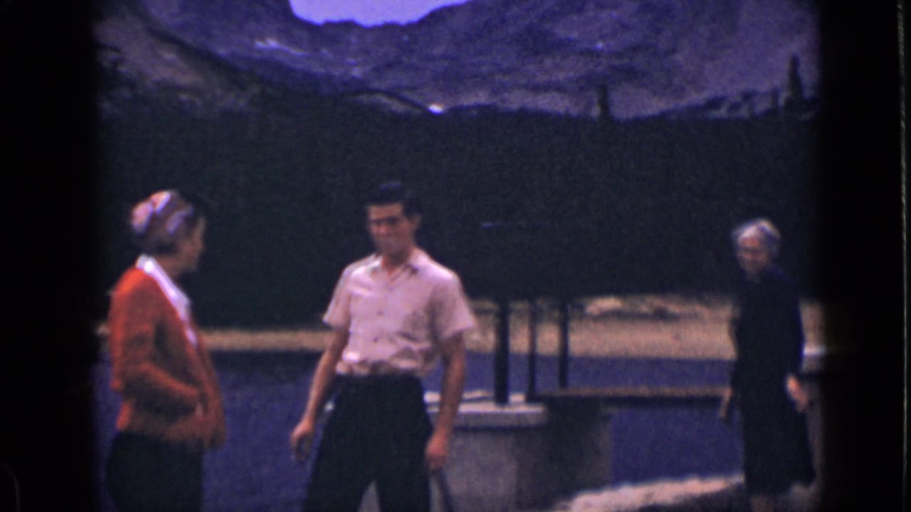 Tourists in 1939 Colorado appear beside a stone bridge railing, with a lake, dark trees, and rugged mountains in the background.