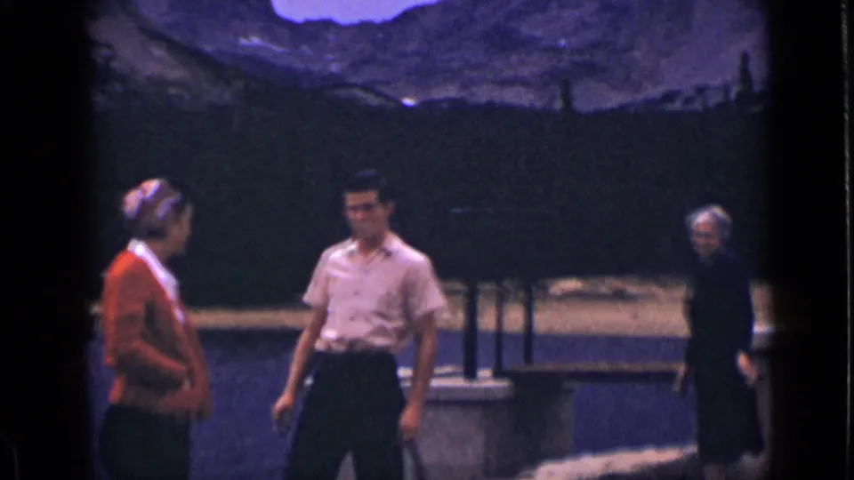 Tourists in 1939 Colorado appear beside a stone bridge railing, with a lake, dark trees, and rugged mountains in the background.