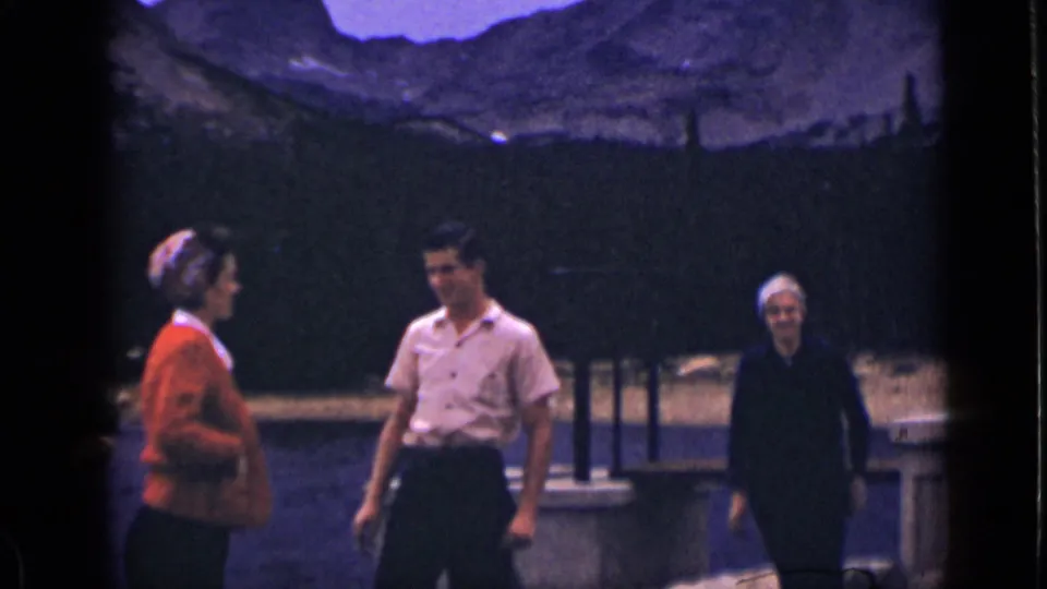 Three adults are visible near the lakeside bridge in Colorado, framed by dark water, evergreen trees, and distant mountain slopes.