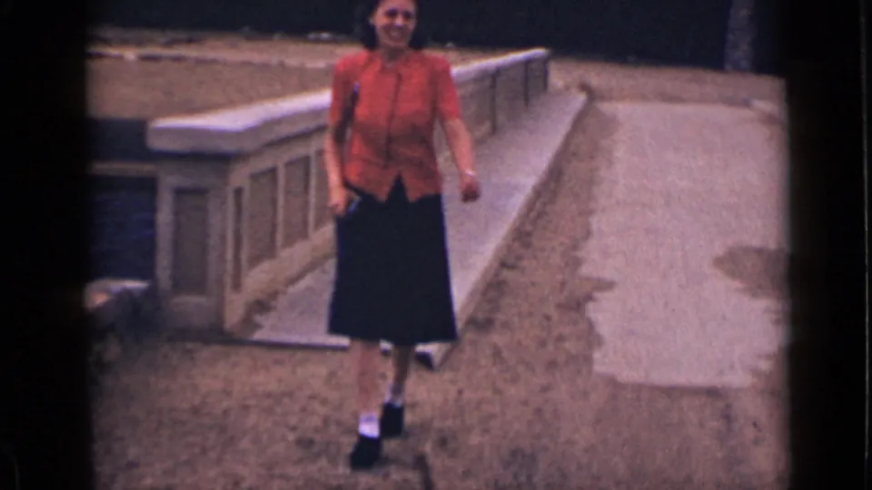 A smiling woman in a red shirt and dark skirt walks toward the camera along the concrete ramp in the 1939 Colorado home movie.