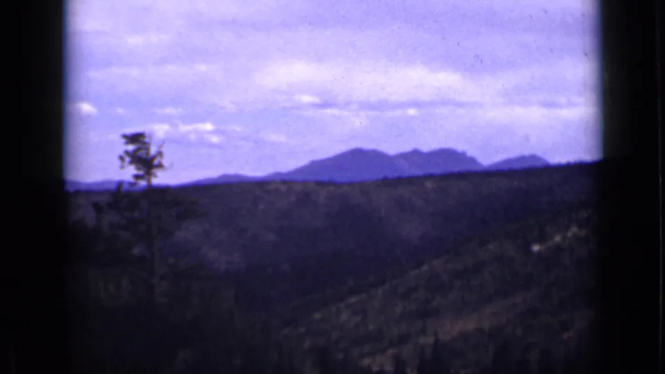 1939 Colorado scenic frame with forested hills, a small pine silhouette, and distant mountains under light clouds.