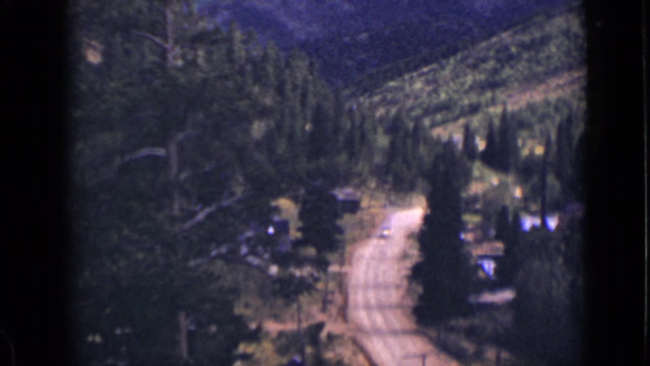 Forested mountain road with cabins or small structures near trees and hillside.