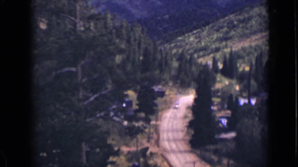 1939 Colorado 8mm frame of a pale dirt road winding through pines below a wooded mountain slope.
