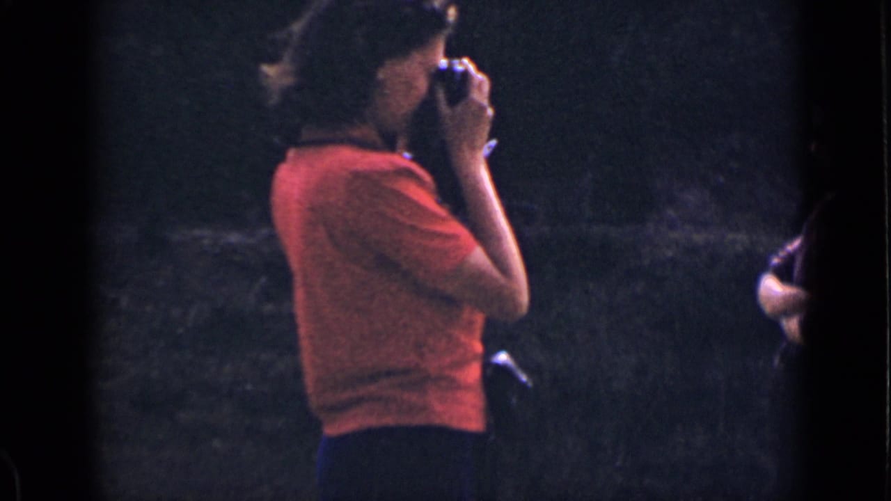 In 1939 Colorado home-movie footage, a woman in a red shirt raises a small camera while standing against a dark wooded background.