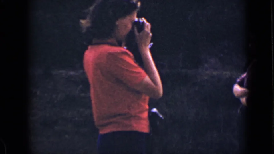 In 1939 Colorado home-movie footage, a woman in a red shirt raises a small camera while standing against a dark wooded background.