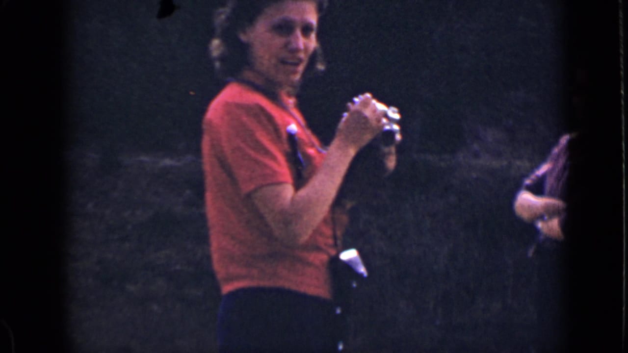 The woman in red turns toward the movie camera while holding her still camera, with a wooded hillside behind her.
