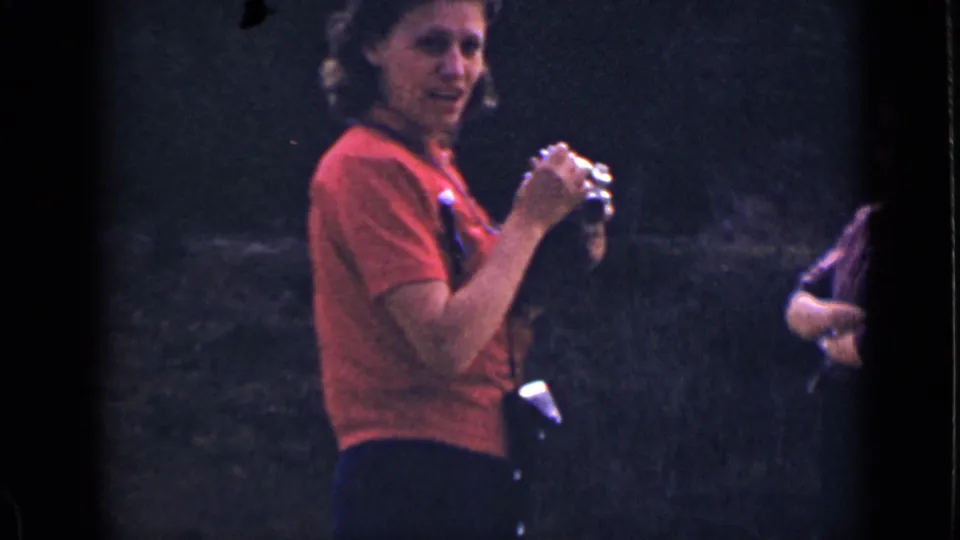 The woman in red turns toward the movie camera while holding her still camera, with a wooded hillside behind her.