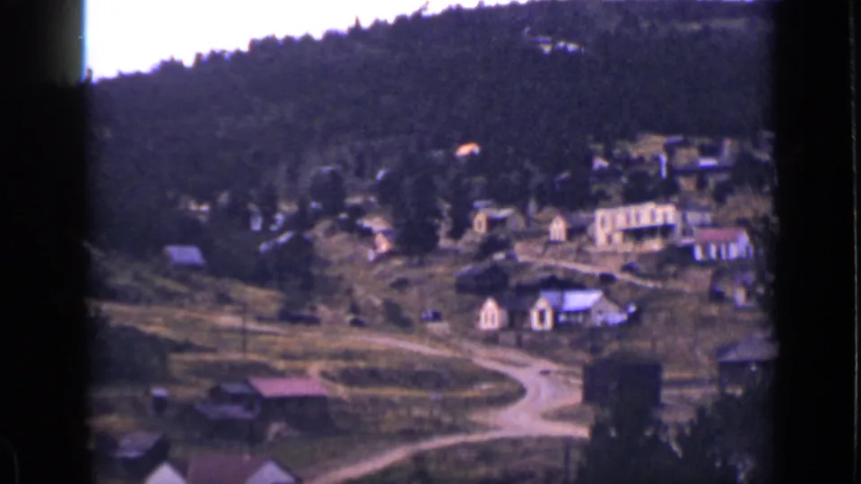 A Colorado mountain village appears from above, with scattered houses, a curving dirt road, and forested slopes beyond.