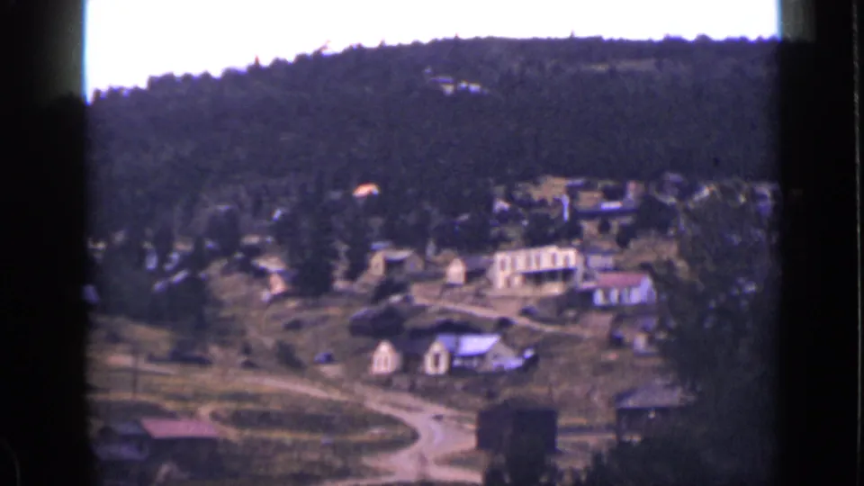 The closing frame holds on the rural Colorado hillside settlement, showing homes, open ground, a road, and dark forest above.