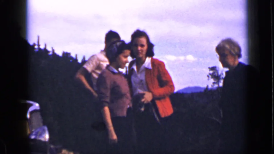 Women in period clothing stand on a Colorado hillside, with a partial car, trees, and distant mountains behind them.