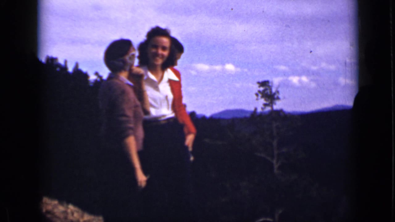 Three women stand on a mountain overlook with distant hills and sky behind them.